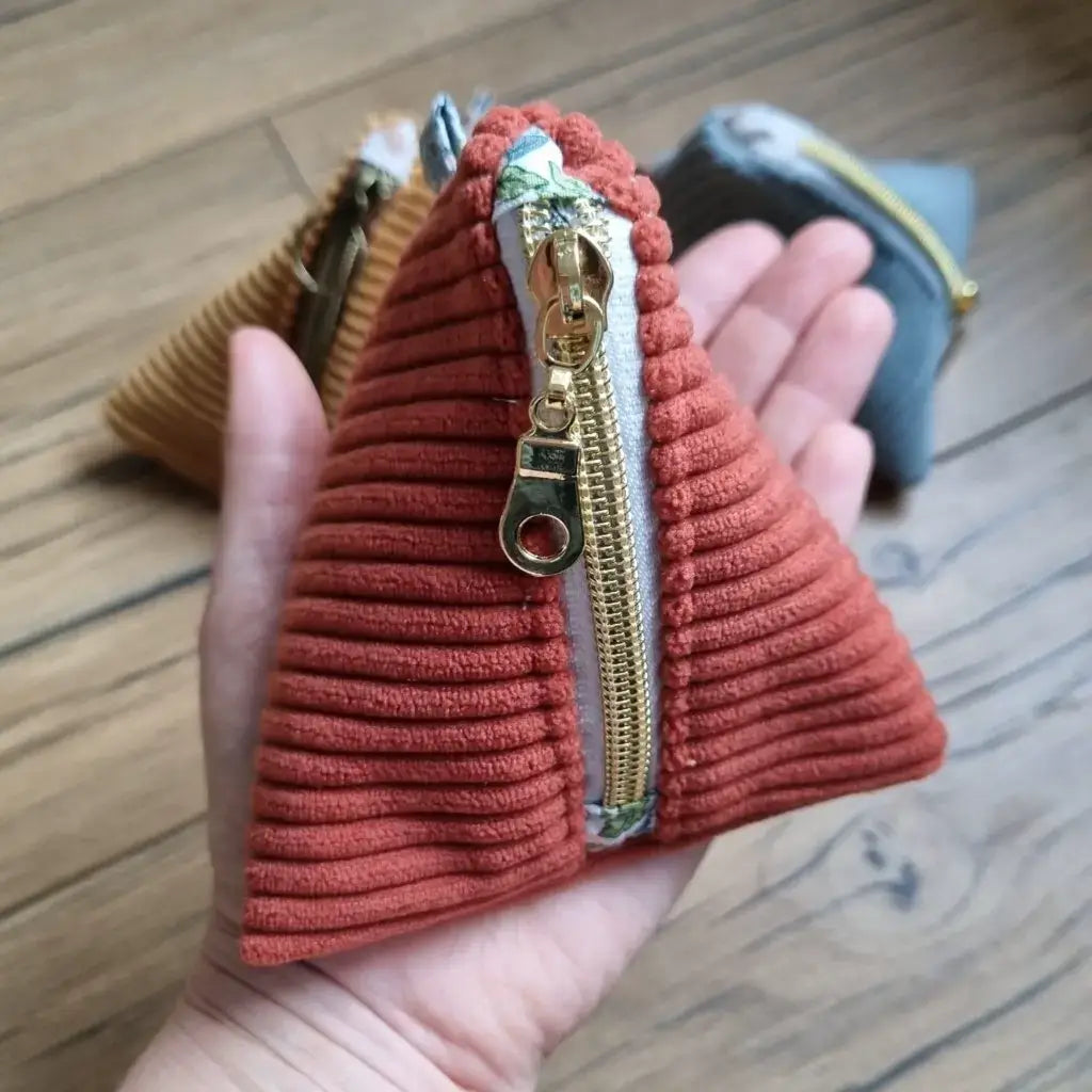 Main soutenant une trousse berlingot en velours côtelé de couleur terracotta. A l'arrière plan, on distingue deux autres trousses berlingots jaune et bleu clair posées au sol sur un parquet en chêne. 