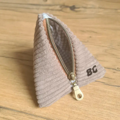 Trousse berlingot en tissu velours côtelé taupe et intérieur carreaux beiges posée sur un parquet en chêne. Ce berlingot est personnalisé avec les initiales "BG" floquées en noir sur l'un de ses côtés.