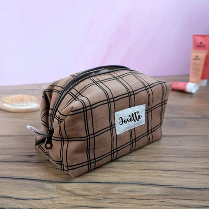 Trousse de toilette carrée en tissu à motif carreaux marron, fermée par une fermeture éclair dorée et personnalisée au prénom “Josette” sur une étiquette en tissu. Accessoire de rangement artisanal posé sur une table en bois, idéal pour le maquillage ou les produits de beauté. Fabrication soignée, au style rétro et chic, parfaite comme idée cadeau personnalisée.