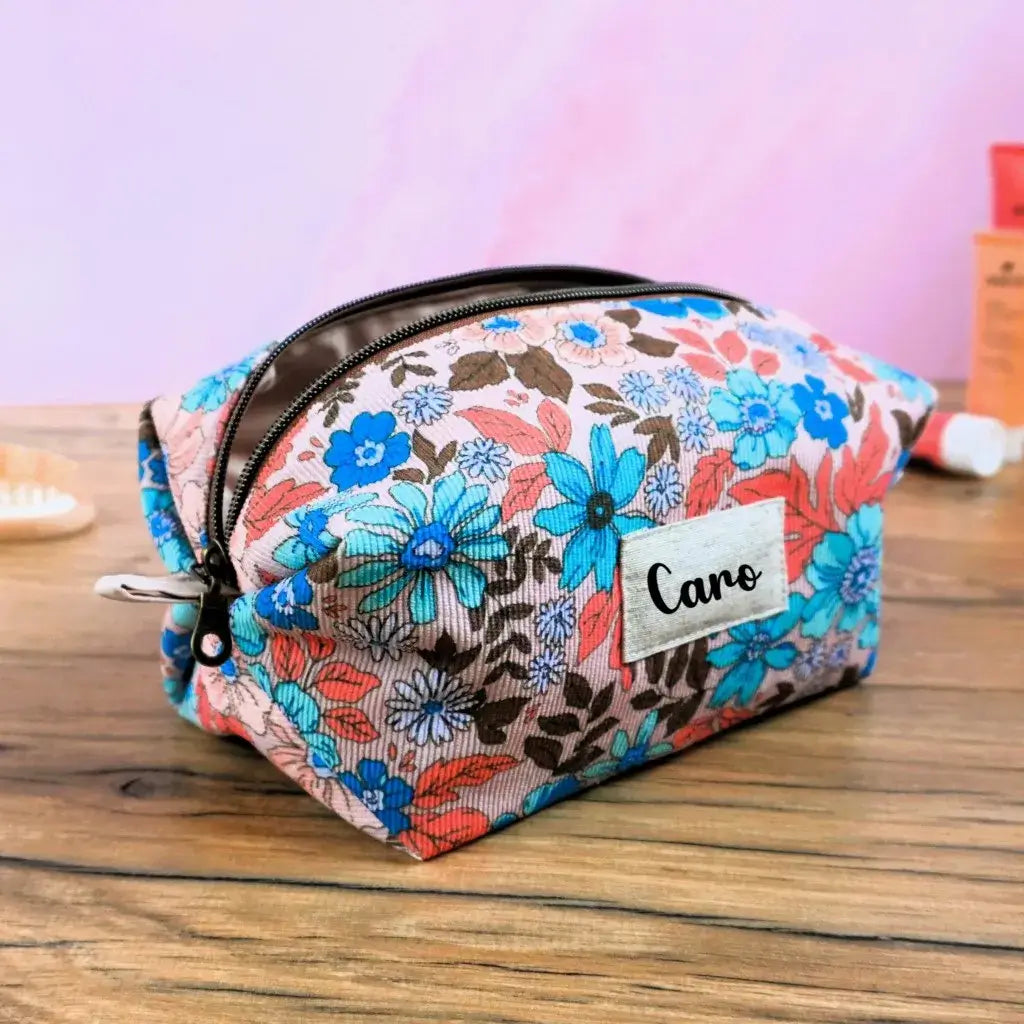 Trousse carrée personnalisée en tissu à motif fleuri vintage, décorée de fleurs turquoise, corail et marron sur fond clair. Fermeture éclair dorée et étiquette en tissu floquée du prénom “Caro”. Accessoire de rangement fait main idéal pour le maquillage, les soins ou les voyages. Design coloré et féminin, parfait comme cadeau personnalisé artisanal fabriqué en France.