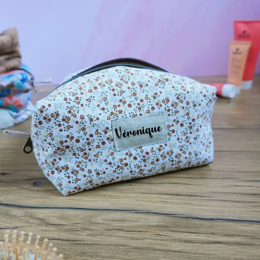 Trousse carrée personnalisée en tissu fleuri à petites fleurs orange et marron sur fond écru, ornée d’une étiquette en tissu avec le prénom “Véronique”. Accessoire de rangement artisanal pratique pour le maquillage, les soins ou les bijoux. Finition soignée, style champêtre et délicat, parfait comme idée cadeau personnalisée et fabriquée à la main.