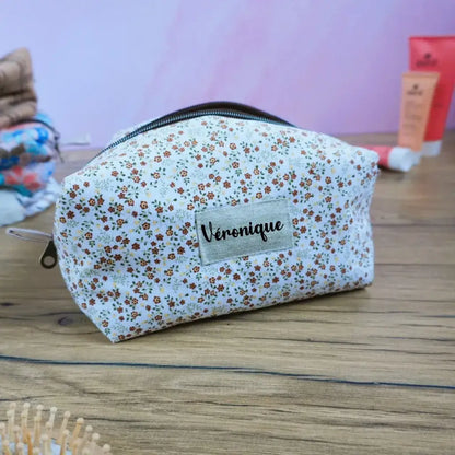 Trousse carrée personnalisée en tissu fleuri à petites fleurs orange et marron sur fond écru, ornée d’une étiquette en tissu avec le prénom “Véronique”. Accessoire de rangement artisanal pratique pour le maquillage, les soins ou les bijoux. Finition soignée, style champêtre et délicat, parfait comme idée cadeau personnalisée et fabriquée à la main.