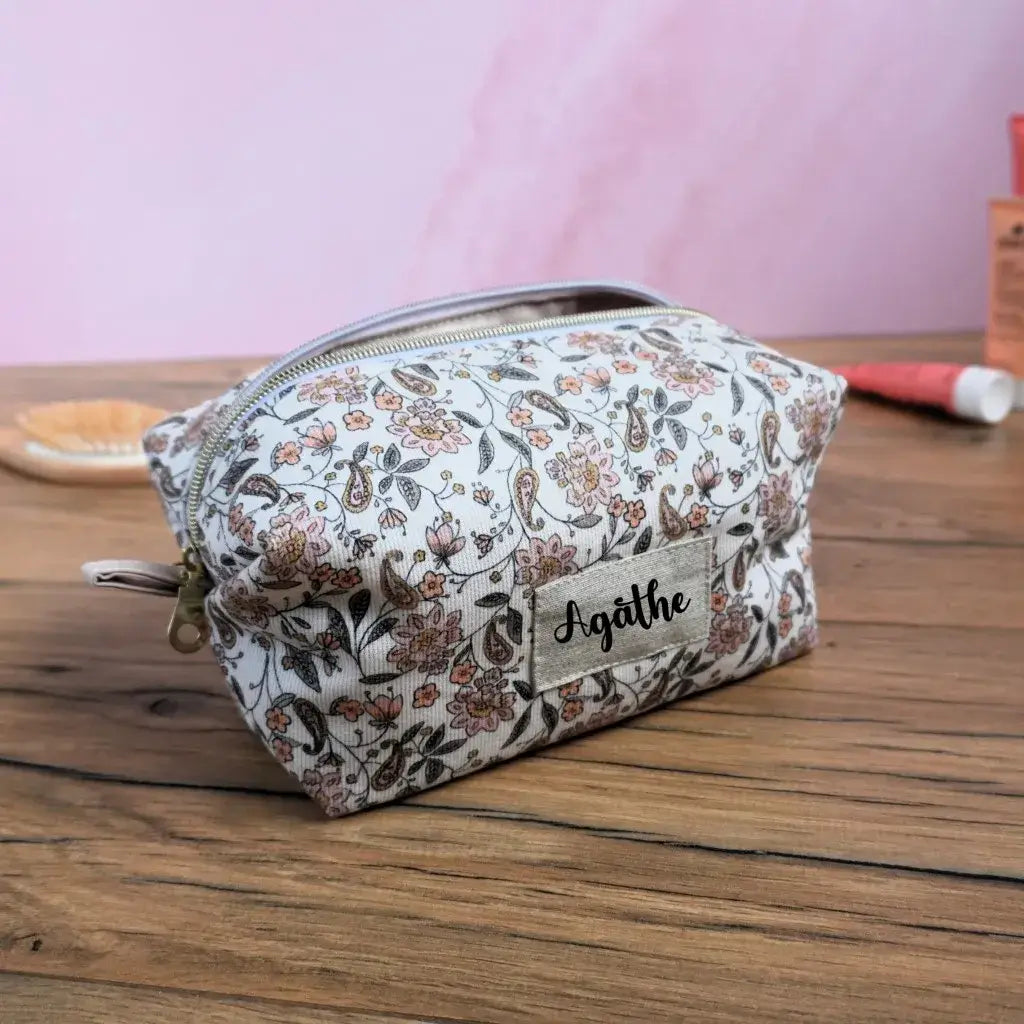 Trousse carrée personnalisée en tissu à motif fleuri délicat, mêlant nuances de beige, rose poudré et brun sur fond clair. Fermeture éclair dorée et étiquette en lin beige floquée du prénom “Agathe”. Accessoire de rangement artisanal idéal pour le maquillage, les accessoires de voyage ou les petits objets du quotidien. Élégante, féminine et faite main en France.