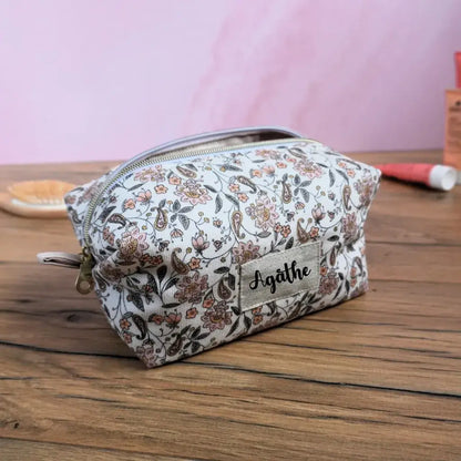 Trousse carrée personnalisée en tissu à motif fleuri délicat, mêlant nuances de beige, rose poudré et brun sur fond clair. Fermeture éclair dorée et étiquette en lin beige floquée du prénom “Agathe”. Accessoire de rangement artisanal idéal pour le maquillage, les accessoires de voyage ou les petits objets du quotidien. Élégante, féminine et faite main en France.