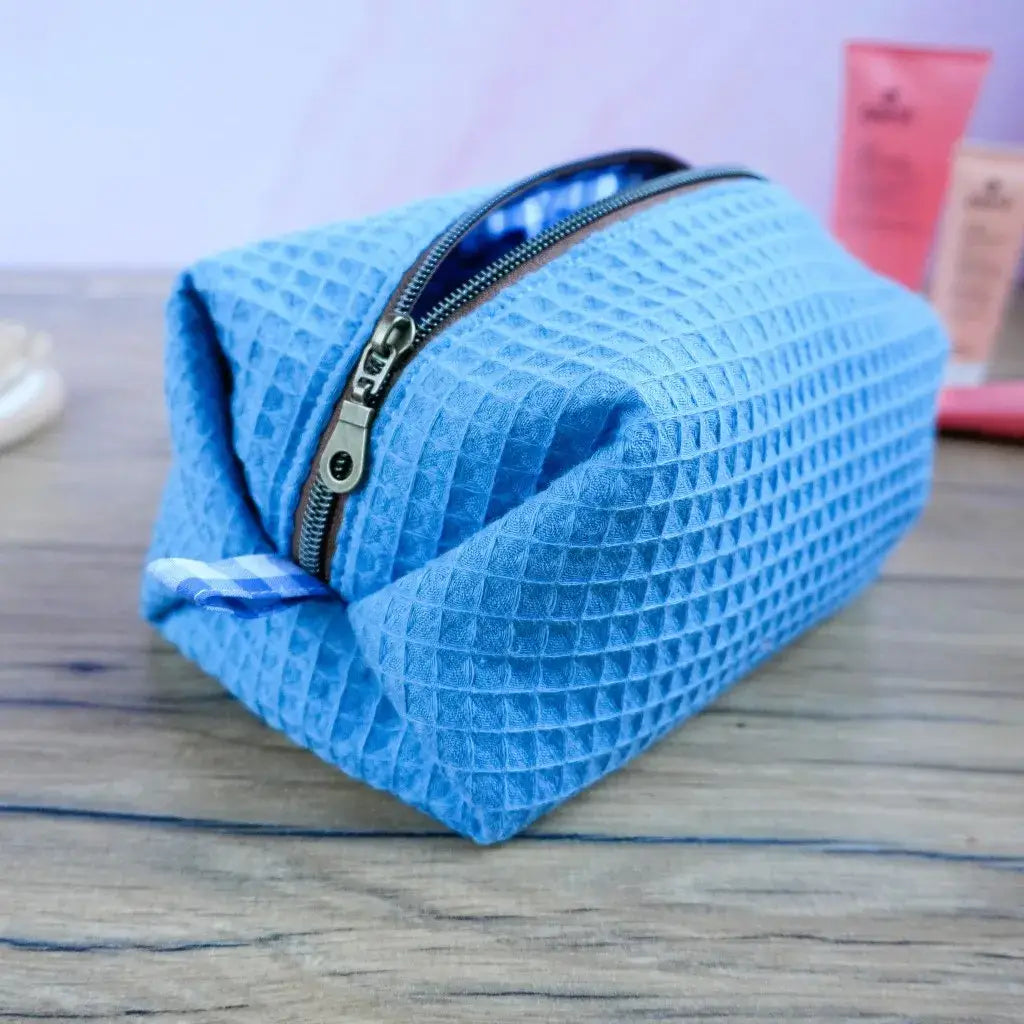 Trousse de toilette carrée en tissu nid d’abeille bleu denim posée sur une table en bois, fermée par une fermeture éclair métallique argentée. Accessoire de voyage artisanal fait main, idéal pour ranger maquillage, produits de soin ou accessoires de bain. Doublure à motif vichy bleu, design moderne et pratique, parfait pour homme ou femme. Idée cadeau personnalisable et tendance pour le quotidien ou les vacances.