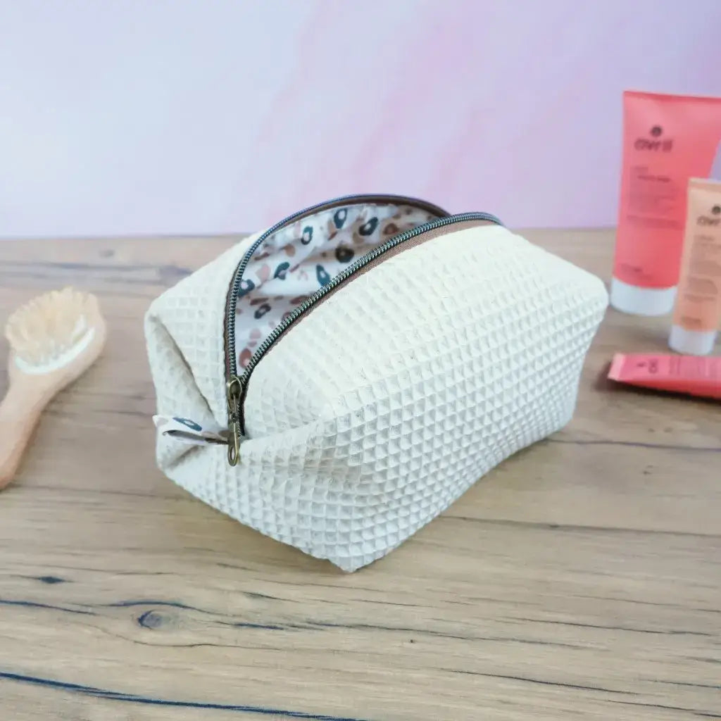 Trousse de toilette carrée en tissu nid d’abeille beige naturel, posée sur un fond en bois avec accessoires de beauté en arrière-plan. Fermeture éclair en métal bronze et doublure à motif léopard discret. Fabrication artisanale soignée, idéale pour le rangement des produits de soin, maquillage ou accessoires de voyage. Design intemporel et raffiné, parfait pour offrir ou compléter une routine bien-être élégante.