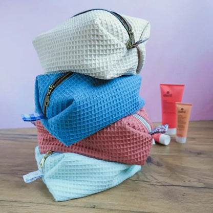 Empilement de quatre trousses carrées en tissu nid d’abeille, déclinées en coloris naturel, bleu denim, marsala et vert menthe. Disposées sur une table en bois avec produits de soin en arrière-plan, elles illustrent une collection artisanale élégante et coordonnée. Chaque trousse présente une fermeture éclair métallique et une doublure imprimée unique. Accessoires faits main parfaits pour le rangement, les voyages ou un coffret cadeau harmonieux.
