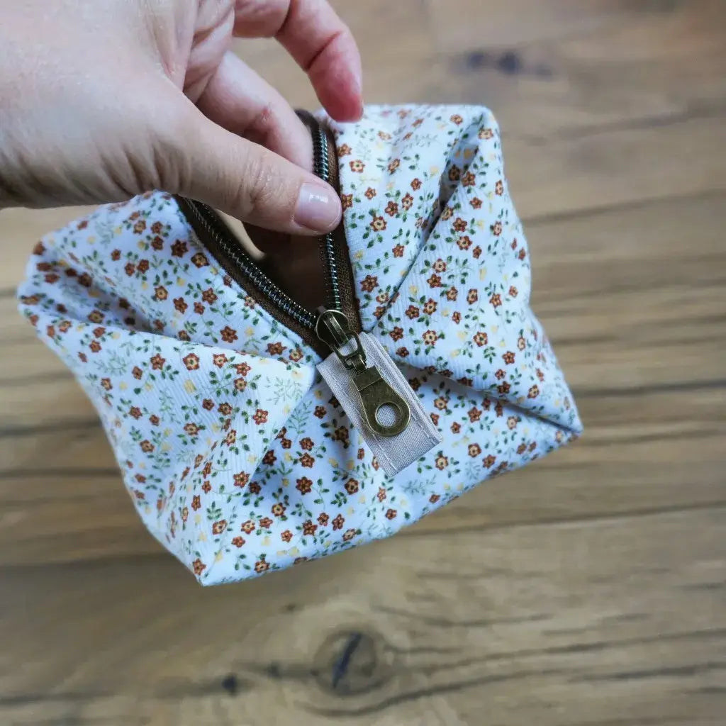 Vue rapprochée d’une trousse carrée en tissu fleuri blanc et orangé, tenue en main pour montrer la fermeture éclair en métal bronze et la finition couture soignée. Accessoire artisanal personnalisé, pratique pour le rangement du maquillage ou des accessoires de voyage, au style champêtre et élégant.