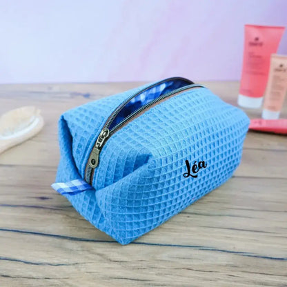 Trousse de toilette carrée personnalisée en tissu nid d’abeille bleu denim avec le prénom “Léa” floqué sur le devant. Modèle artisanal fait main, doublé d’un tissu vichy bleu et blanc et fermé par une fermeture éclair métallique. Posée sur une table en bois avec accessoires de beauté en arrière-plan, cette trousse au style doux et moderne allie praticité et élégance. Idéale comme cadeau unique ou accessoire de voyage personnalisé.