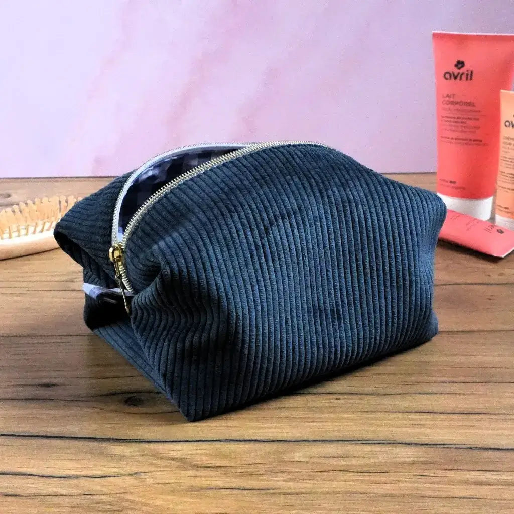 Trousse carrée en velours côtelé bleu foncé posée sur une table en bois, entourée de produits de beauté. Sa fermeture dorée et sa texture douce allient élégance et praticité. Grâce à sa forme stable et sa grande capacité, elle permet de ranger maquillage, soins ou accessoires de toilette. Un accessoire artisanal chic et personnalisable, parfait pour un usage quotidien.