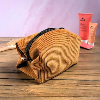 Trousse carrée en velours côtelé couleur jaune posée sur une table en bois, accompagnée de produits de beauté. Sa fermeture éclair marron contrastante souligne son design moderne. Pratique et spacieuse, elle permet de ranger facilement maquillage, soins ou accessoires. Confectionnée à la main, cette trousse personnalisable allie douceur du velours et élégance artisanale.