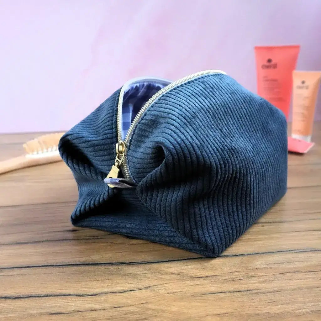 Trousse carrée en velours côtelé bleu marine avec fermeture éclair dorée, présentée sur une table en bois dans un univers de salle de bain. Sa forme souple et sa large ouverture assurent un accès pratique aux produits de beauté ou accessoires de voyage. Finition artisanale et doublure en coton à motifs pour un accessoire élégant, durable et personnalisable.