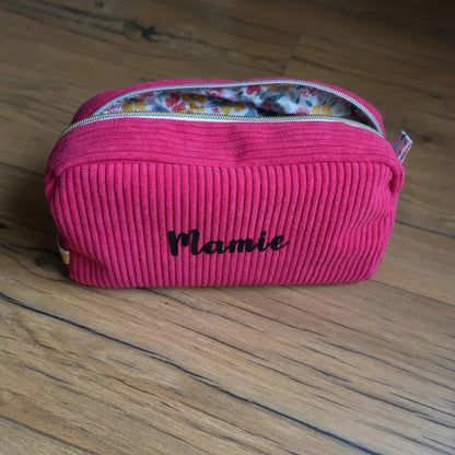 Trousse de toilette en velours côtelé framboise, personnalisée avec l’inscription “Mamie” floquée en velours noir sur le devant. Elle est équipée d’une fermeture éclair argentée et doublée d’un tissu intérieur à motifs floraux colorés. Posée sur un sol en bois, cette trousse artisanale allie praticité et élégance, idéale pour ranger produits de beauté, accessoires ou effets personnels lors des déplacements.