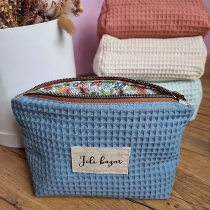 Au premier plan, une trousse zippée en tissu nid d'abeille de coloris bleu denim personnalisée avec l'inscription "joli bazar" floquée sur une étiquette en toile. A l'arrière-plan, trois autres trousses similaires de coloris naturel, marsala et menthe sont empilées les unes sur les autres à côté d'un bouquet de fleurs séchées.
