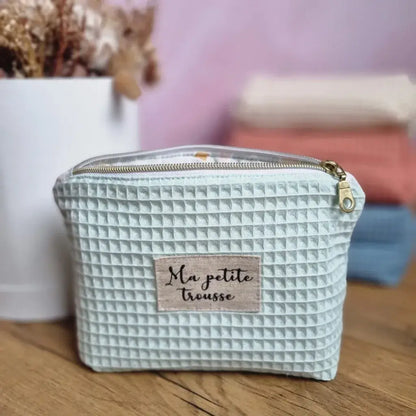 Au premier plan, une trousse zippée en tissu nid d'abeille de coloris menthe personnalisée avec l'inscription "Ma petite trousse" floquée sur une étiquette en toile. A l'arrière-plan, trois autres trousses similaires de coloris naturel, marsala et bleu denim sont empilées les unes sur les autres à côté d'un bouquet de fleurs séchées.