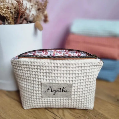 Au premier plan, une trousse zippée en tissu nid d'abeille de coloris naturel personnalisée avec le prénom "Agathe" floquée sur une étiquette en toile. A l'arrière-plan, trois autres trousses similaires de coloris menthe, marsala et bleu denim sont empilées les unes sur les autres à côté d'un bouquet de fleurs séchées.