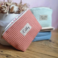 Au premier plan, une trousse en tissu nid d'abeille de coloris marsala et personnalisée avec le prénom "Cathy" floqué sur une étiquette en toile. Cette trousse est posée de biais devant un bouquet de fleurs séchées. A l'arrière plan, on distingue trois autres trousses similaires empilées les unes sur les autres et de coloris menthe, naturel et bleu denim.