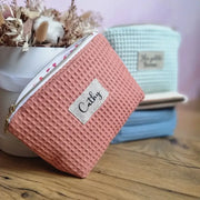 Au premier plan, une trousse en tissu nid d'abeille de coloris marsala et personnalisée avec le prénom "Cathy" floqué sur une étiquette en toile. Cette trousse est posée de biais devant un bouquet de fleurs séchées. A l'arrière plan, on distingue trois autres trousses similaires empilées les unes sur les autres et de coloris menthe, naturel et bleu denim.