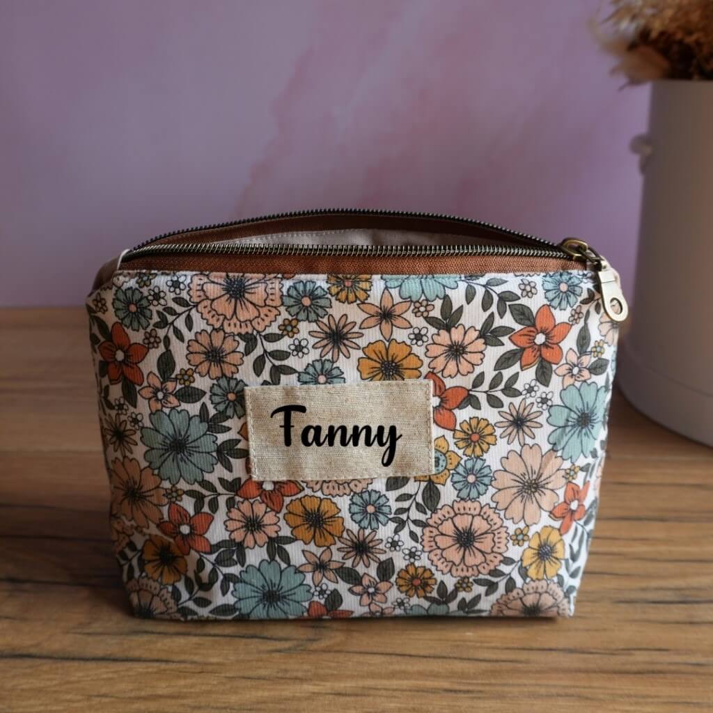 Trousse zippée personnalisée en tissu fleuri rétro aux tons pastel et orangés, décorée d’une étiquette en lin avec le prénom Fanny floqué en noir. Accessoire artisanal idéal comme trousse de toilette, pochette à maquillage ou rangement pour le sac à main. Son motif floral élégant et sa personnalisation en font un cadeau pratique et original pour femme, ado ou enfant.