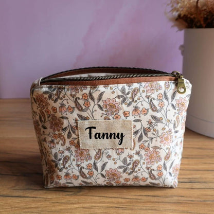 Trousse zippée personnalisée en tissu clair à motif floral délicat dans des nuances de beige, brun et corail, ornée d’une étiquette en lin avec le prénom Fanny floqué en noir. Pochette artisanale élégante et fonctionnelle, parfaite pour ranger maquillage, fournitures ou accessoires. Son imprimé raffiné et sa personnalisation en font un accessoire pratique et esthétique, idéal au quotidien ou comme cadeau personnalisé.
