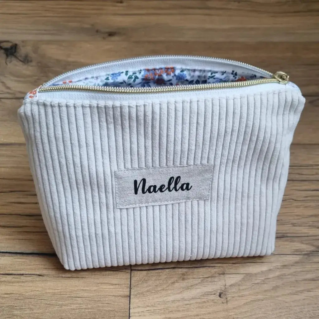 Trousse zippée en velours côtelé beige avec une doublure intérieure en coton imprimé à motif fleuri. Cette trousse dispose d'une étiquette en tissu cousue en façade sur laquelle est floqué le prénom "Naella" en écriture noire.