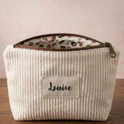 Trousse zippée en velours côtelé beige avec une doublure intérieure en coton imprimé à motif fleuri. Cette trousse dispose d'une étiquette en tissu cousue en façade sur laquelle est floqué le prénom "Naella" en écriture noire.