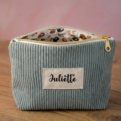 Trousse zippée en velours côtelé bleu clair avec une doublure intérieure en coton imprimé à motif fleuri. Cette trousse dispose d'une étiquette en tissu cousue en façade pouvant accueillir un prénom ou une inscription personnalisée qui sera floquée dessus. A l'arrière plan, on distingue quatre autres trousses zippées en velours côtelé du même type et de coloris divers disposées les unes contre les autres devant une lampe.