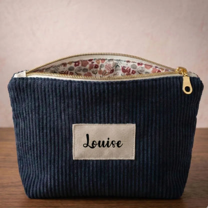 Trousse zippée en velours côtelé bleu foncé avec une doublure intérieure en coton imprimé à motif fleuri. Cette trousse dispose d'une étiquette en tissu cousue en façade pouvant accueillir un prénom ou une inscription personnalisée qui sera floquée dessus. A l'arrière plan, on distingue quatre autres trousses zippées en velours côtelé du même type et de coloris divers disposées les unes contre les autres devant une lampe.