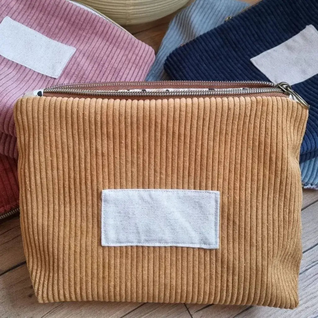 Trousse zippée en velours côtelé jaune. Cette trousse dispose d'une étiquette en tissu cousue en façade pouvant accueillir un prénom ou une inscription personnalisée qui sera floquée dessus. A l'arrière-plan, on distingue d'autres trousses zippées du même type et de coloris divers entassées les unes sur les autres.