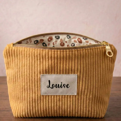 Trousse zippée en velours côtelé jaune. Cette trousse dispose d'une étiquette en tissu cousue en façade pouvant accueillir un prénom ou une inscription personnalisée qui sera floquée dessus. A l'arrière-plan, on distingue d'autres trousses zippées du même type et de coloris divers entassées les unes sur les autres.