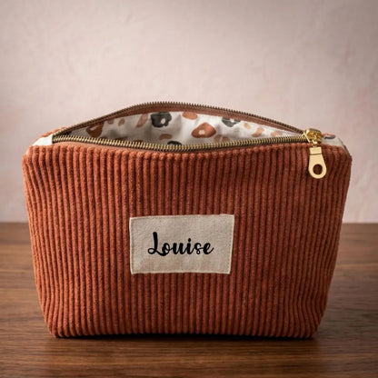 Trousse zippée en velours côtelé marron avec une doublure intérieure en coton imprimé à motif fleuri. Cette trousse dispose d'une étiquette en tissu cousue en façade pouvant accueillir un prénom ou une inscription personnalisée qui sera floquée dessus.