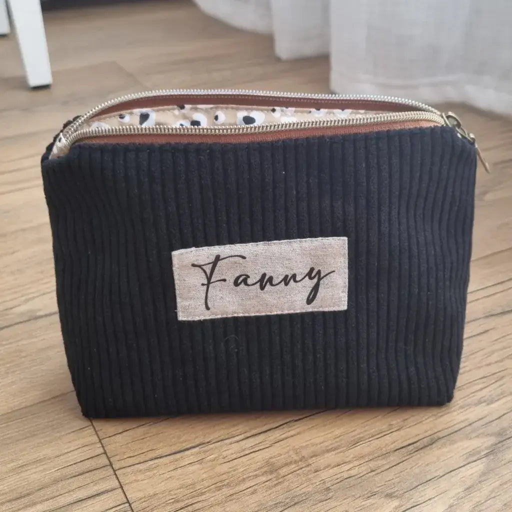 Trousse zippée en velours côtelé noir avec une doublure intérieure en coton imprimé à motif léopard. Cette trousse dispose d'une étiquette en tissu cousue en façade sur laquelle est floqué le prénom "Fanny" en écriture noire.