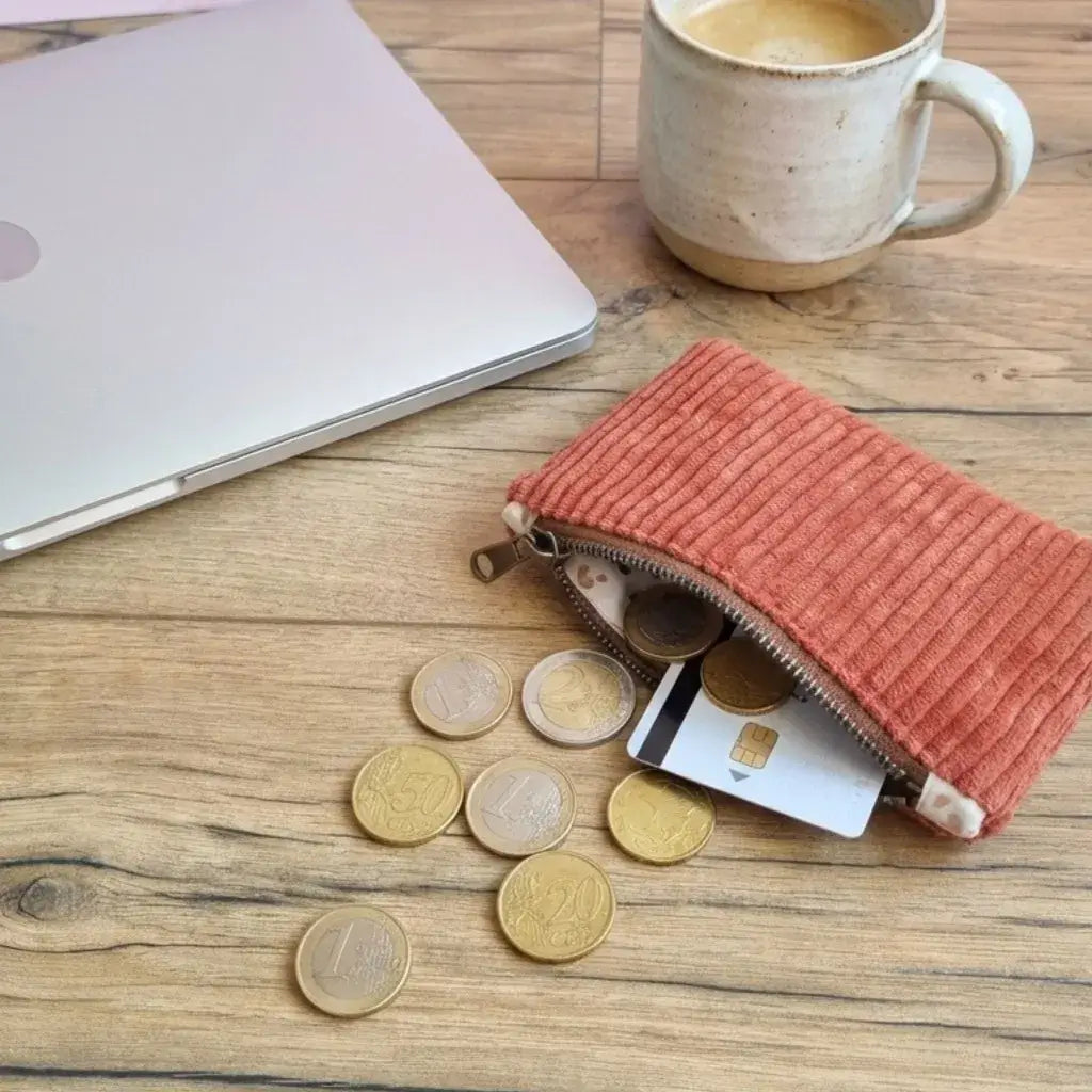 Porte-monnaie en velours côtelé terracotta avec pièces et carte bancaire sur un bureau en bois.