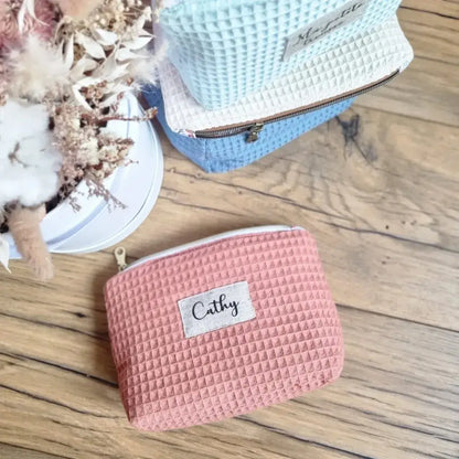 Une trousse zippée en tissu nid d'abeille de coloris marsala est déposée au sol sur un parquet en chêne. Cette trousse est personnalisée avec le prénom "Cathy". Dans le coin supérieur gauche, vu de dessus, un bouquet de fleurs séchées. A droite de ce bouquet, trois autres trousses en tissu nid d'abeille de coloris menthe, naturel et bleu denim sont empilées les une sur les autres.