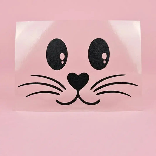 Flex thermocollant noir : visage de lapin avec yeux ovales, nez en cœur et moustaches, sur support transparent et fond rose.