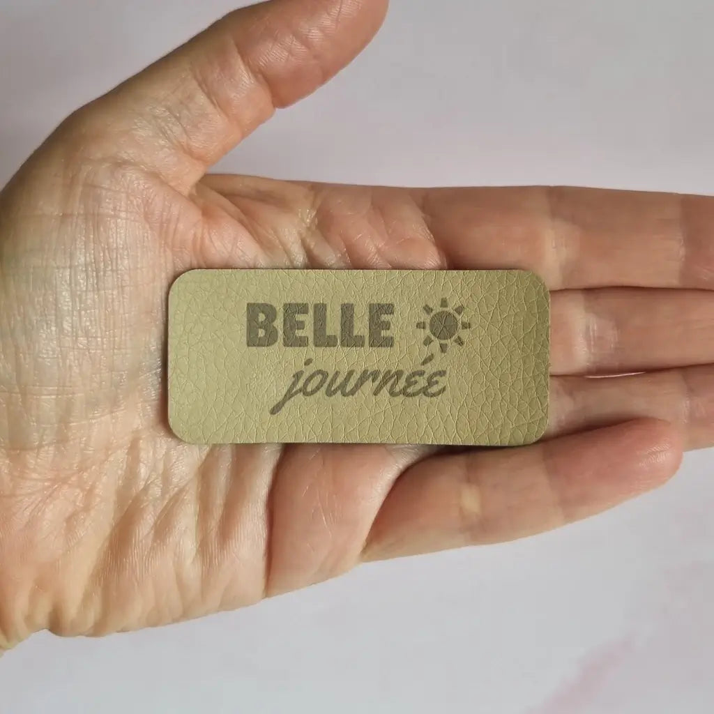 Étiquette simili cuir gravée Belle journée avec motif soleil, posée au creux de la main