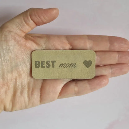 Étiquette simili cuir vert gravée Best mom avec cœur, présentée dans la paume d'une main