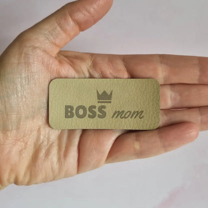 Étiquette simili cuir gravée Boss mom surmontée d'une couronne, vue de face dans la main