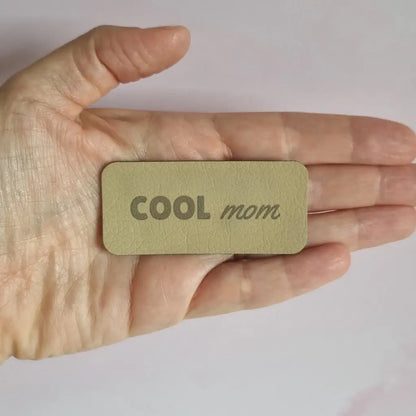 Étiquette à coudre en simili cuir gravée Cool mom, sans motif, posée dans la main
