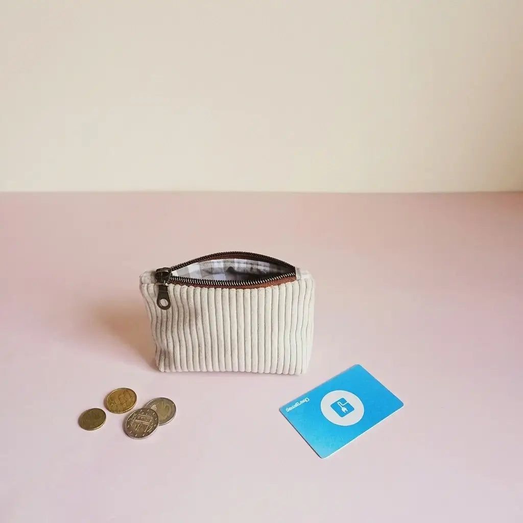 Porte-monnaie zippé écru en velours côtelé avec zip foncé, posé près de pièces de monnaie