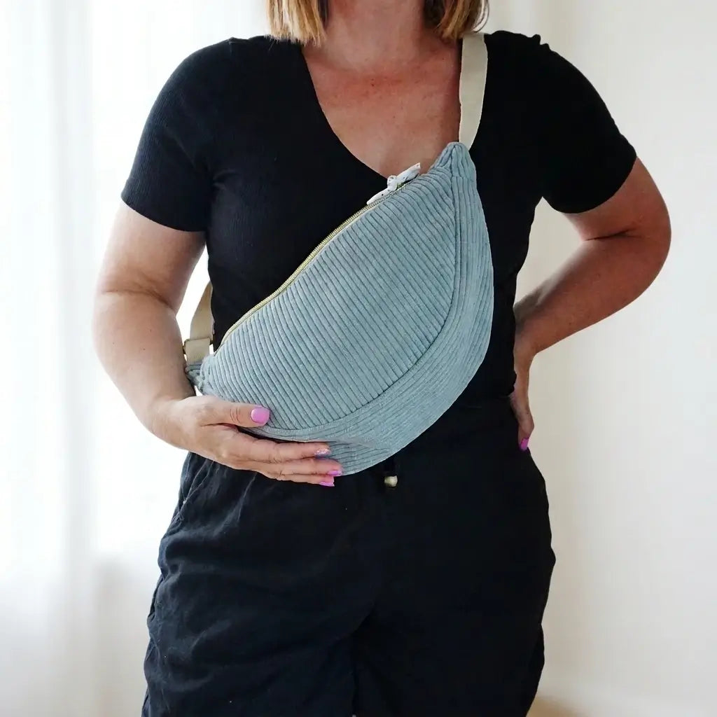 Sac banane demi-lune en velours côtelé bleu clair porté en bandoulière sur une tenue noire