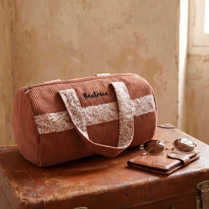 sac polochon velours côtelé rose posé sur valise en cuir style voyage