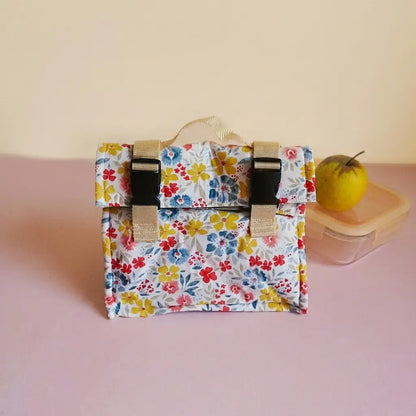 Sac repas à motif fleuri jaune, bleu et rouge, rabat à clips, posé près d'une boîte tupperware et d'une pomme
