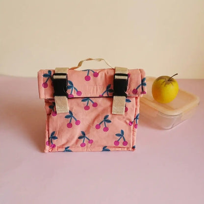 Sac repas rose à motif cerises, poignée beige et fermeture par clips, à côté d'une boîte repas