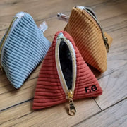 Trois trousses berlingot en tissu velours côtelé posée au sol sur un parquet en chêne. Celle au premier plan est de couleur terracotta, a son zip ouvert et présente des initiales floquées sur le coin inférieur droit. A l'arrière plan, à droite une trousse berlingot jaune, à gauche une trousse berlingot bleu clair.